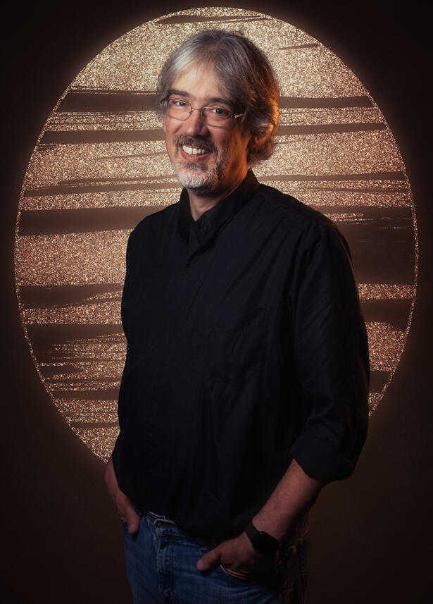 Image of James Wyatt—a middle-aged white man with gray hair and glasses—wearing a black shirt and blue jeans in front of a golden glowing background. Photo by KP11 Studios.