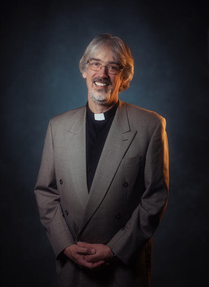 Image of James Wyatt—a middle-aged white man with gray hair and glasses—wearing a clerical collar, black shirt, and gray suit coat in front of a blue background. Photo by KP11 Studios.